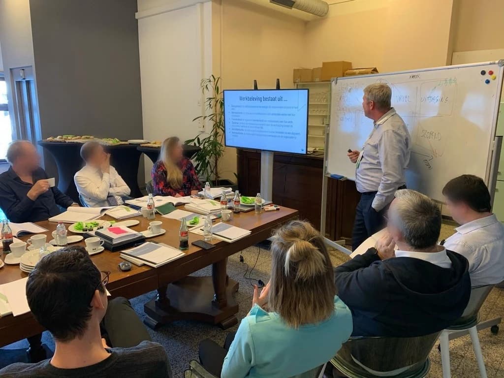Kris Rayen aan het whiteboard tijdens een werksessie met een team; op het scherm naast hem staat 'Werkbeleving bestaat uit'.