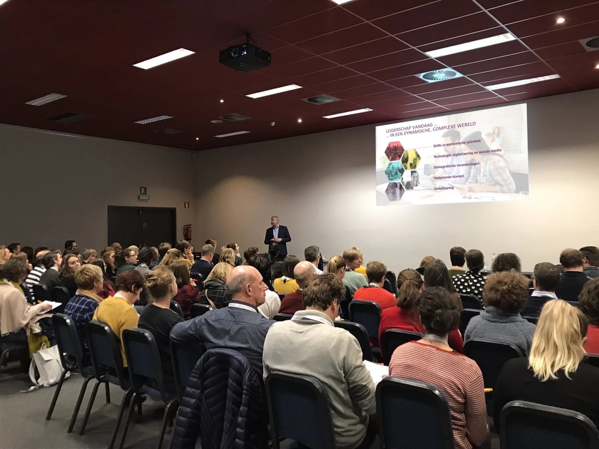 Kris Rayen speaking to a full room, seen from the back; the screen behind him shows a slide on leadership in a dynamic, complex world.